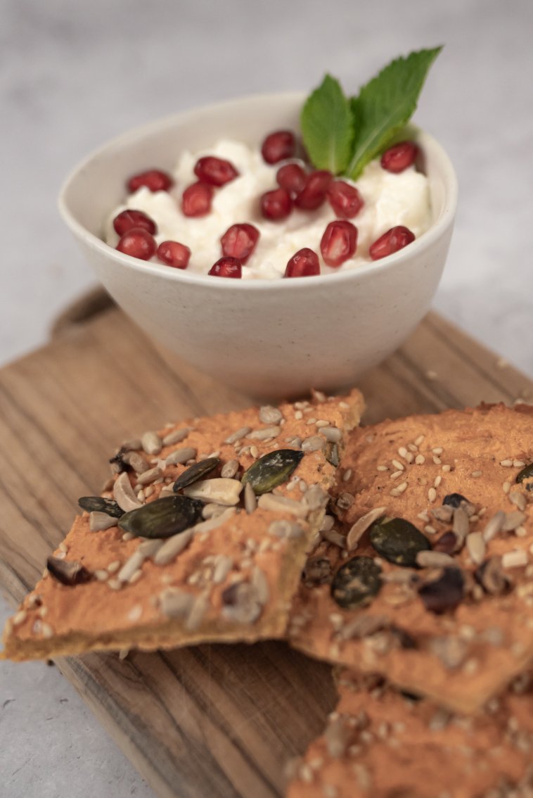 Cracker auf einem Holzbrett, dahinter steht eine Schale mit Joghurt, Granatapfelkernen und Minze.
