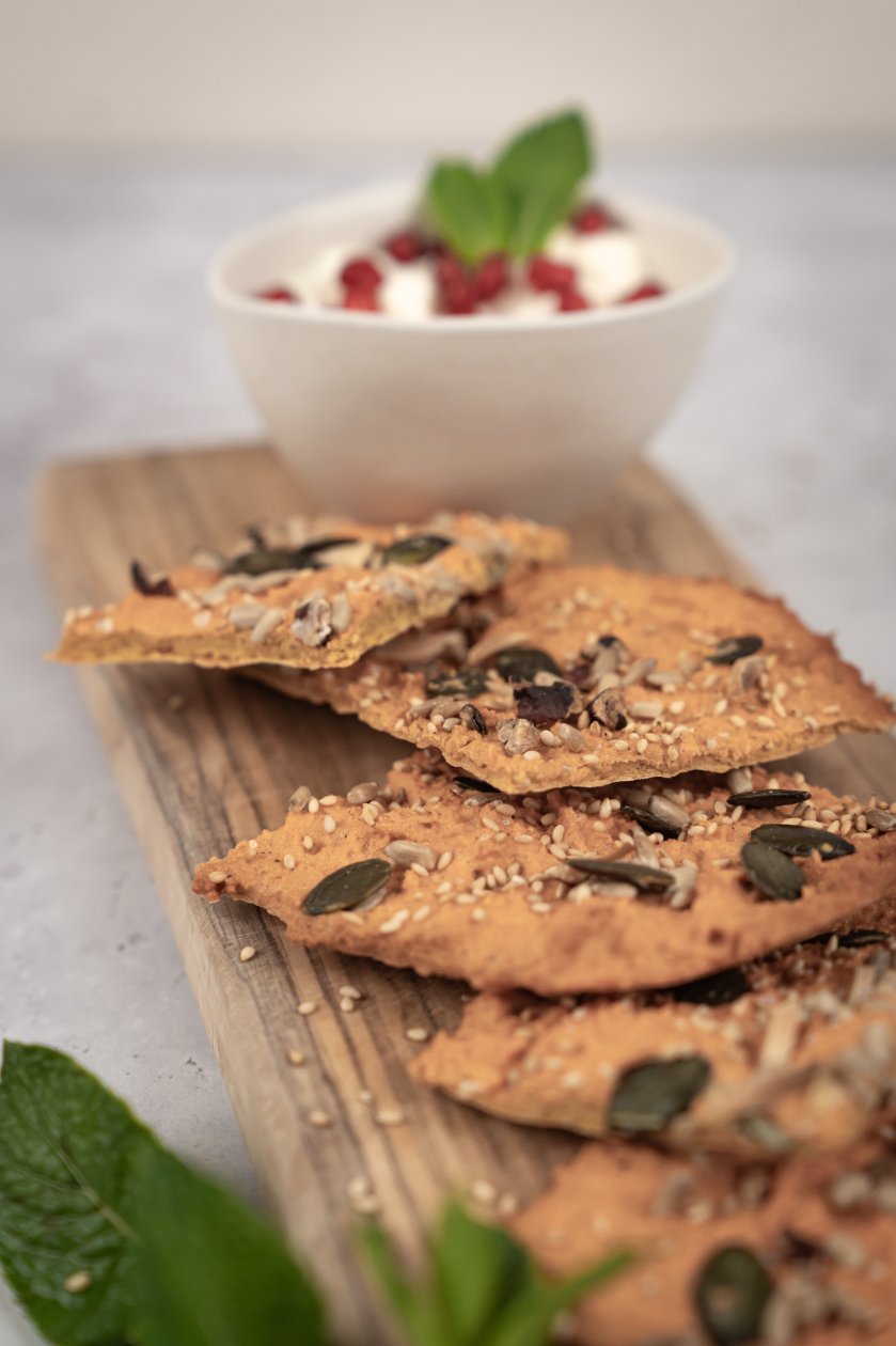 Cracker auf einem Holzbrett, dahinter steht eine Schale mit Joghurt, Granatapfelkernen und Minze.