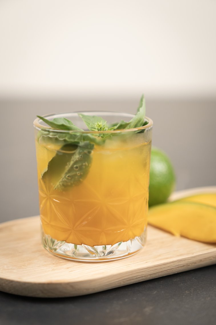 Glas mit dem Drink, garniert mit frischen Kräutern, daneben liegen eine Limette und Mango Stücke.