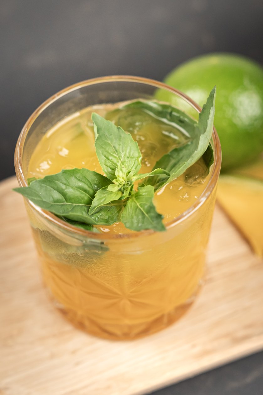 Glas mit dem Drink, garniert mit frischen Kräutern, daneben liegen eine Limette und Mango Stücke.